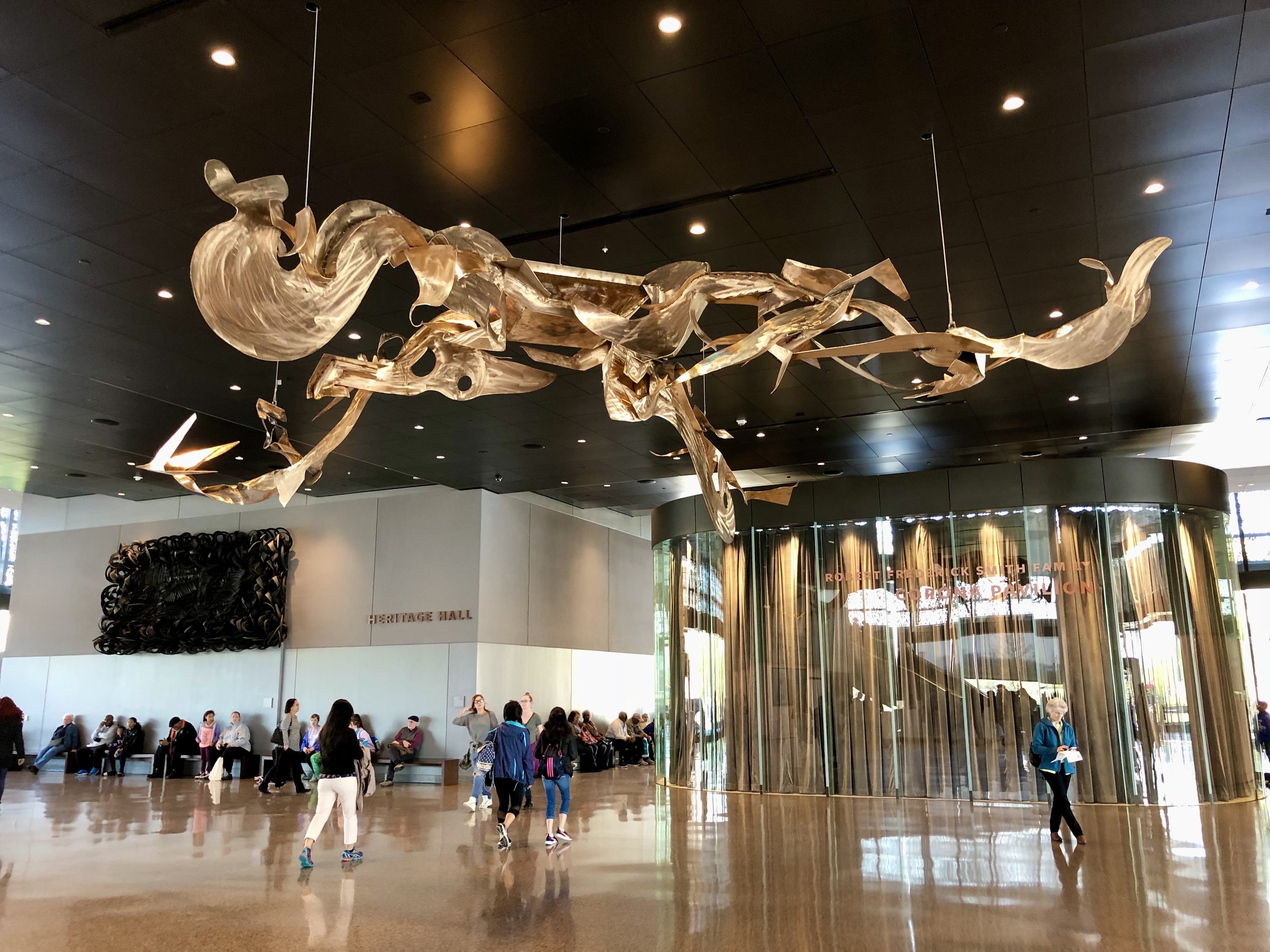 Gold bronze metal that stretches across the ceiling of a building hanging from wires. The sculpture consists of flat, arcing shapes and winglike forms. 