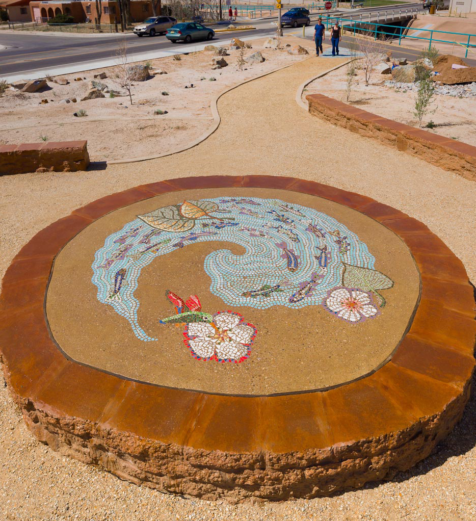 Stone circle in center of park with ceramic white flowers with a hummingbird feeding on nectar and a river in a circle filled with fish.