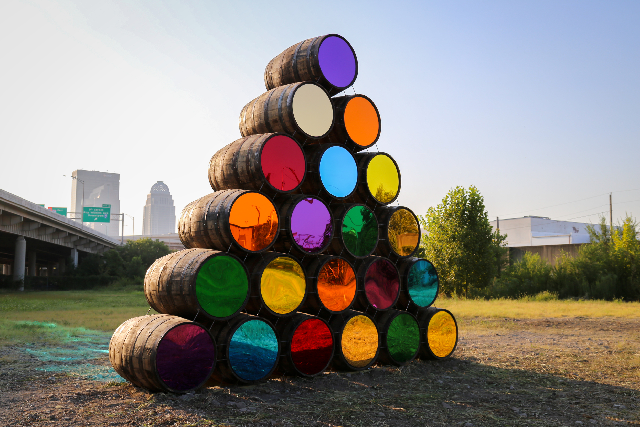 Twenty colorful barrels are placed on top of one another, forming a pyramid outside in the city.