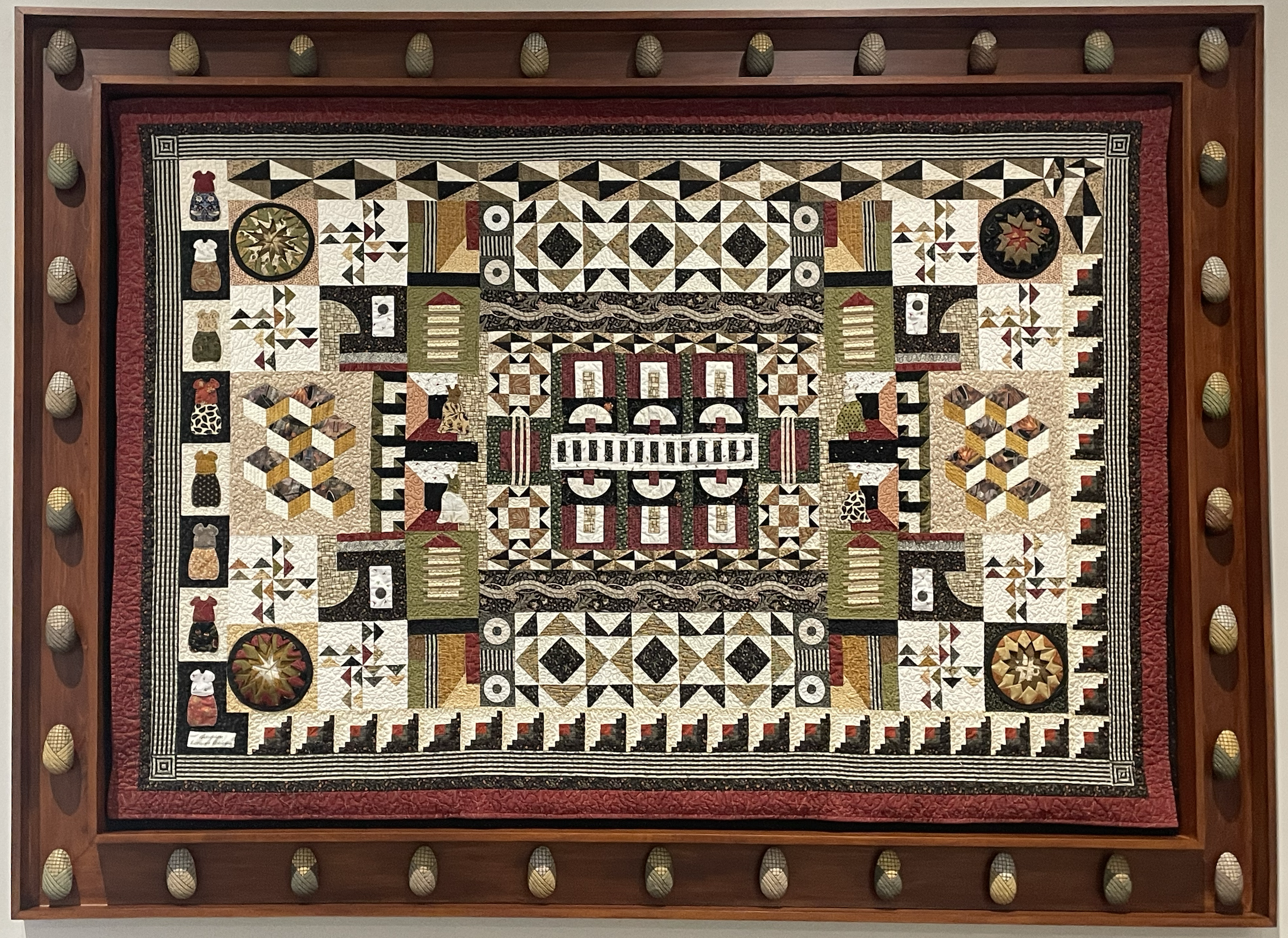 The image shows a quilt framed in a dark wood frame with decorative accents. The quilt is made of various fabrics in black, white, brown, beige, and green, and features a complex geometric pattern. The quilt is displayed against a light-colored wall. The frame is decorated with small, rounded objects that are evenly spaced around the edge.