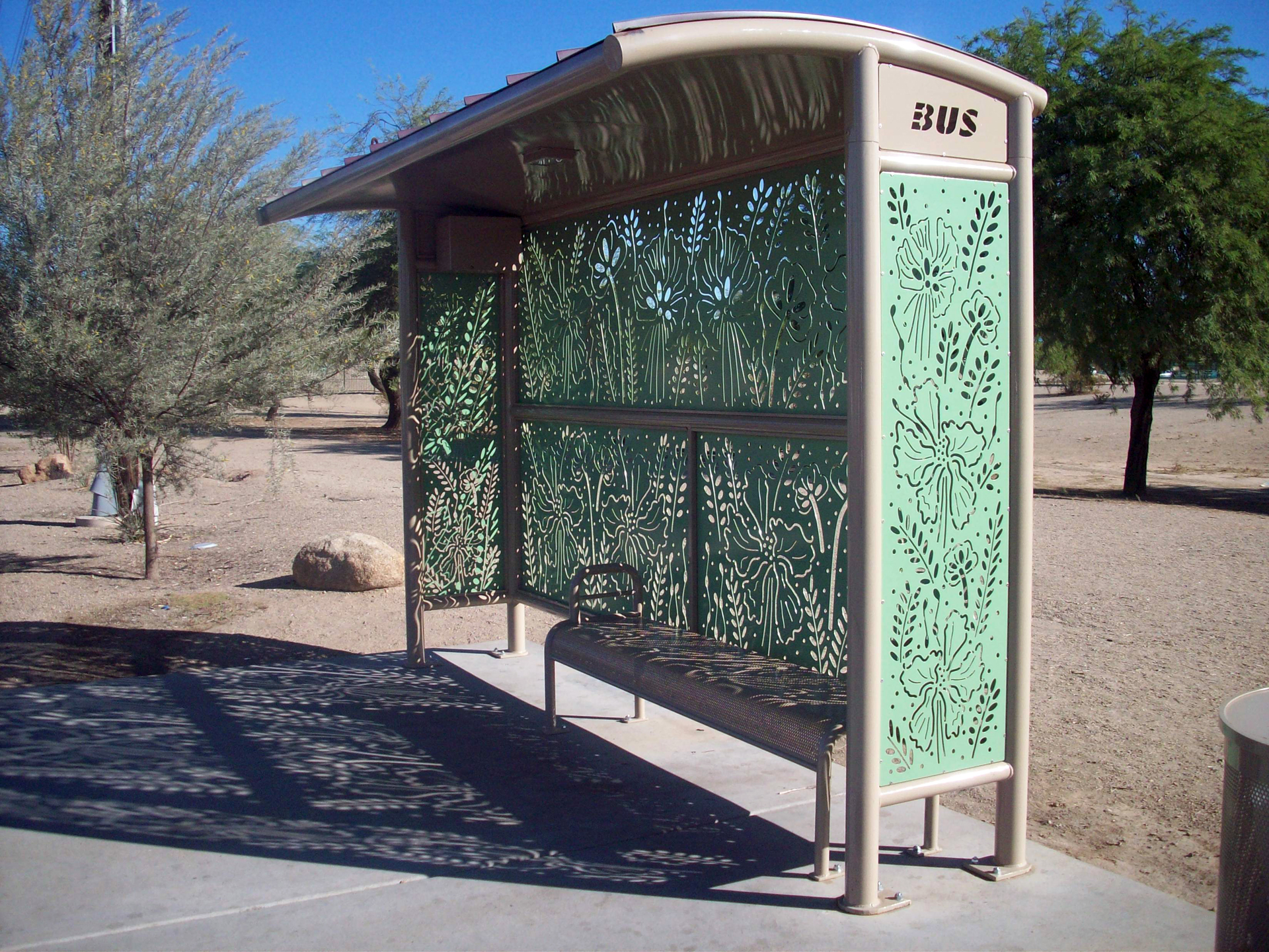 Image of light gray bus shelter with a desertscape behind it. Bus shelter walls are made of cut metal with floral pattern in a light green color.