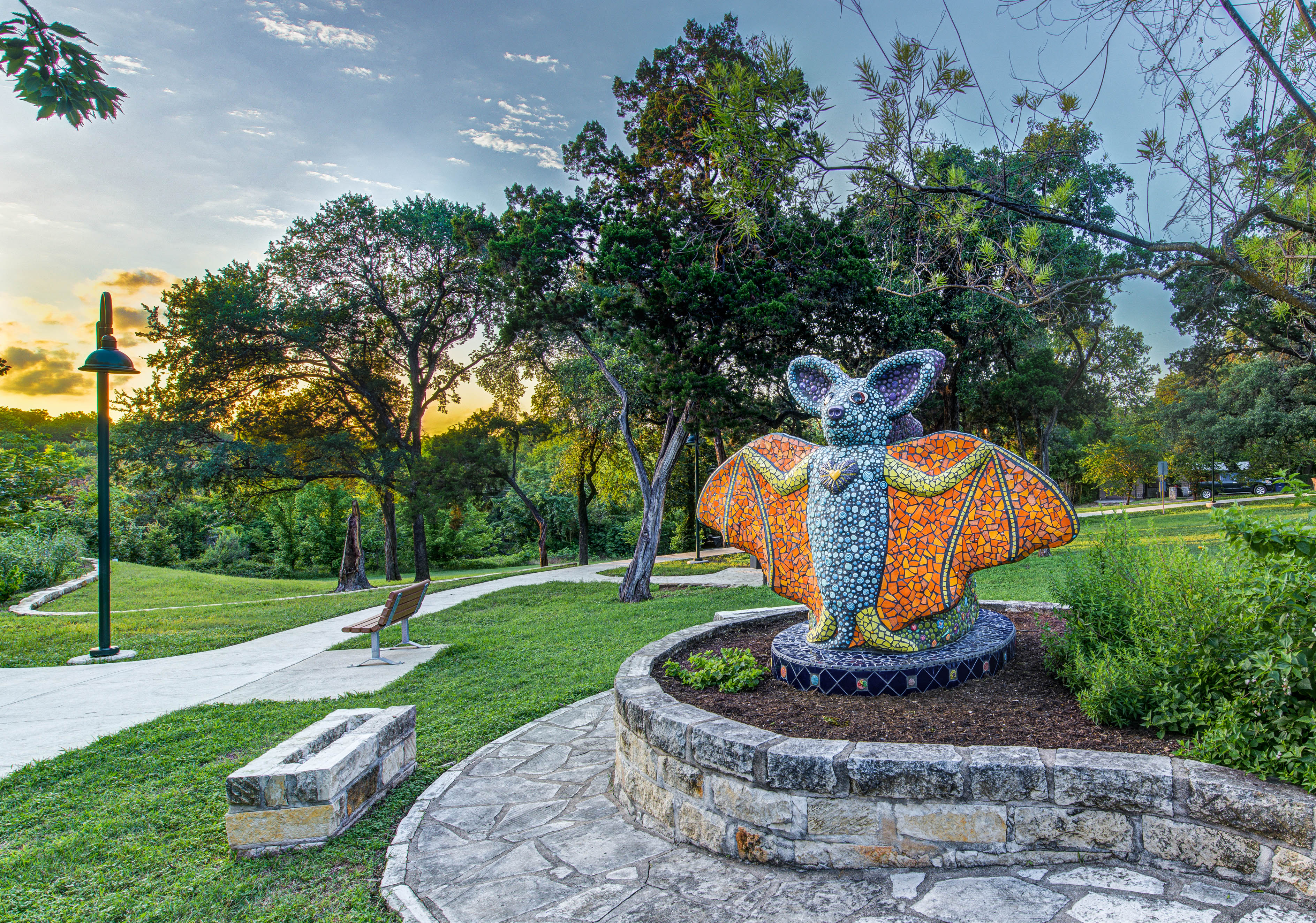 Mosiac bat statue with orange wings and a light and dark blue body standing in a garden with its wings out. 