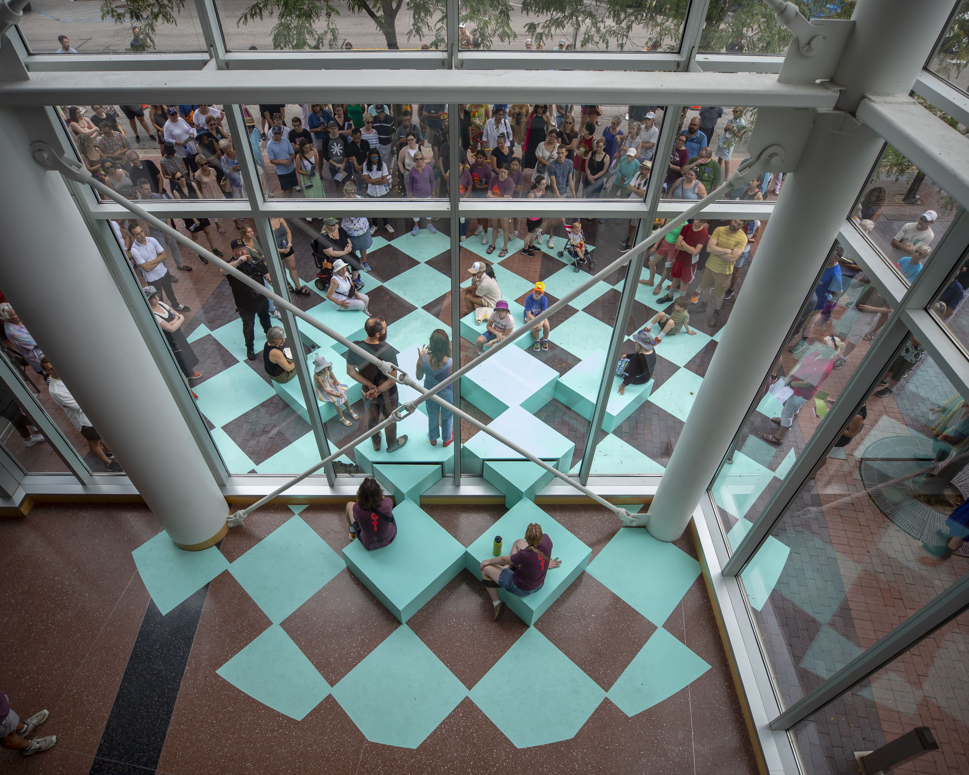 The image shows an indoor space with a large window overlooking an outdoor area. The outdoor space features a checkered pattern of light blue squares, creating a visual effect similar to a giant chessboard. Some of the squares are elevated into platforms, which people are sitting on. There are also a large number of people gathered outside the window, seemingly observing what's happening below. The indoor space appears to be a lobby or atrium, with a patterned floor of brown and light blue tiles. The image is taken from a high vantage point, looking down at the scene.