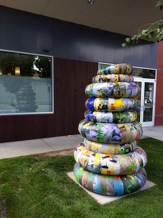 The image shows a large stack of colorful, circular objects, each covered in a different pattern. The objects appear to be made of a flexible material like rubber or plastic and are stacked on top of each other in a conical shape. They are located on a patch of green grass in front of a building.