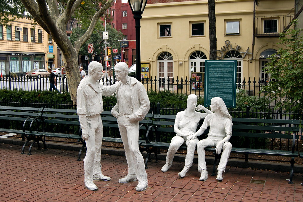 White stone statues of people. Two men stand in front of a bench with one man's arm on the shoulder. Two women sit on the bench with one woman's hand on the other's leg. 