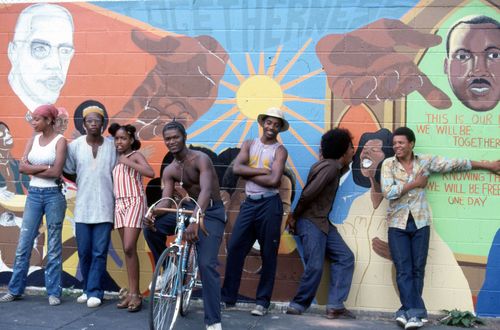 A collective of people stands together, smiling in front of a colorful mural, highlighting community and artistic expression.