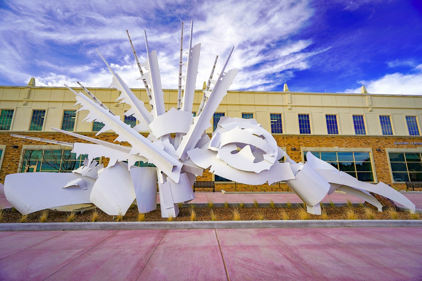 White large metal sculpture similar to a fan with sharp ends in front of building. 